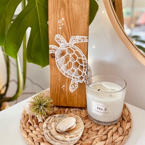 May include: A wooden sign featuring a white painted sea turtle design. The sign is displayed on a table with a woven placemat, a small plant, and a candle labelled "No. 3 Pomegranate".