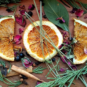 May include: A wooden surface with dried orange slices, cinnamon sticks, cloves, juniper berries, and rose petals. The ingredients are arranged in a decorative pattern and are surrounded by green sprigs.