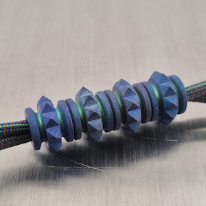 Op de afbeelding: Close-up van een set blauwe metalen kralen met een gestructureerd, geometrisch ontwerp. De kralen zijn geregen op een veelkleurig koord. De kralen hebben een donkerblauwe kleur met groene en paarse accenten.