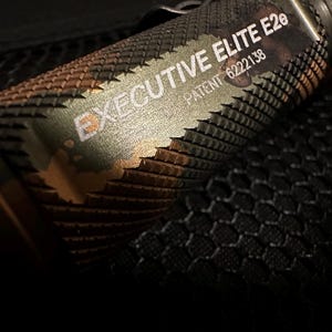 Puede incluir: Primer plano de una linterna con un agarre texturizado y las palabras "EXECUTIVE ELITE E2e" y "PATENT 6222138" impresas en el lateral. La linterna tiene un patr&oacute;n de camuflaje con tonos verdes, marrones y dorados.