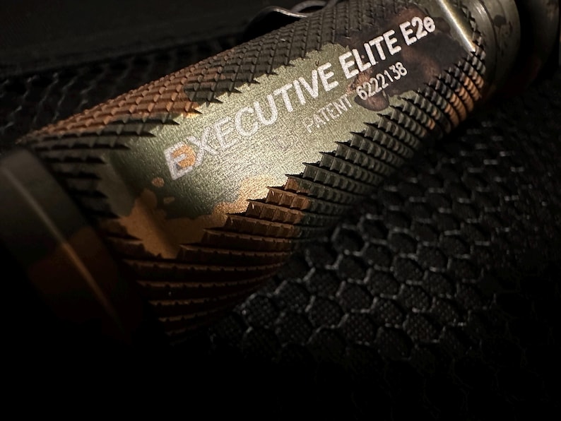 Puede incluir: Primer plano de una linterna con un agarre texturizado y las palabras "EXECUTIVE ELITE E2e" y "PATENT 6222138" impresas en el lateral. La linterna tiene un patr&oacute;n de camuflaje con tonos verdes, marrones y dorados.
