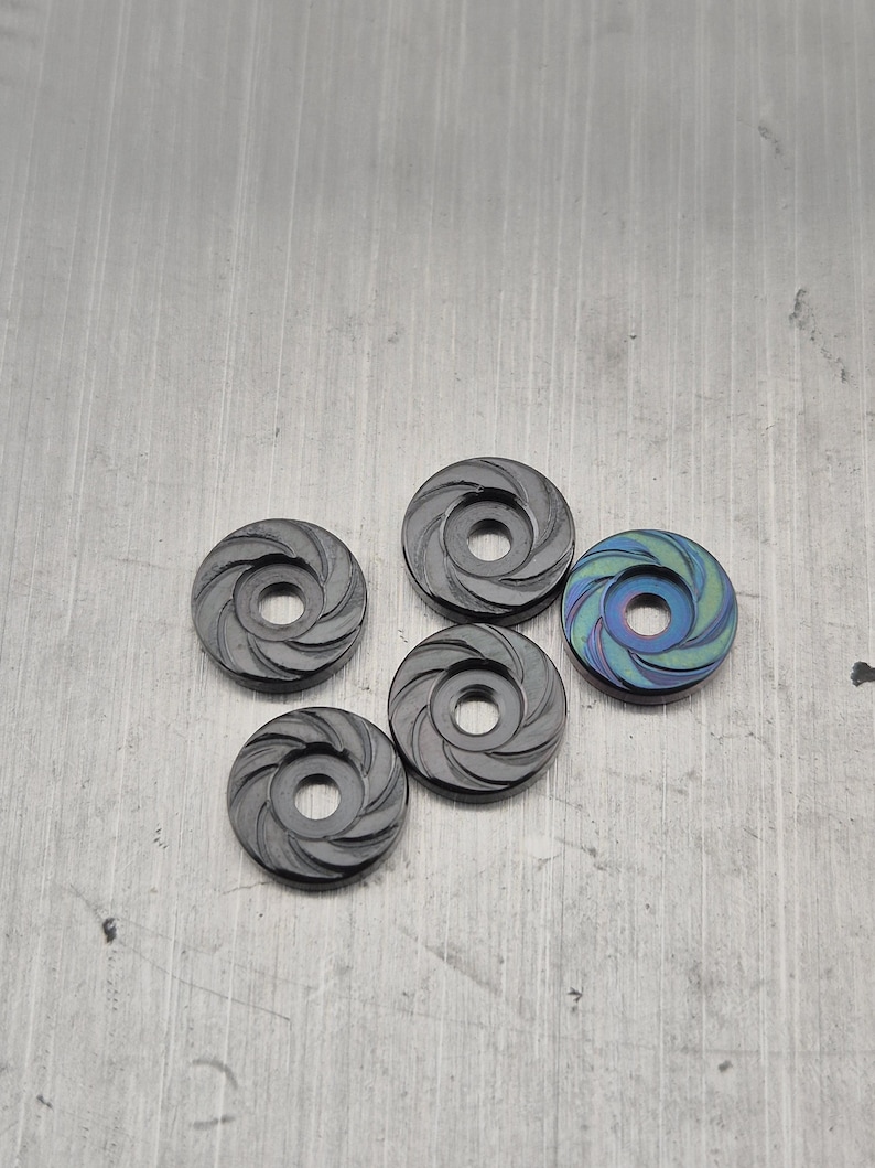 Op de afbeelding: Vijf kleine, cirkelvormige metalen objecten met een centraal gat en een spiraalvormig ontwerp. Vier zijn donkergrijs en &eacute;&eacute;n heeft een blauwgroene iriserende afwerking. Ze liggen op een getextureerd, metalen oppervlak.