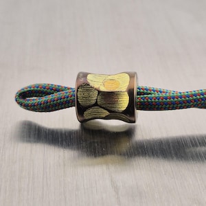 May include: A bronze-colored cylindrical bead with gold-colored abstract designs. The bead is strung on a colorful rope with red, blue, and green threads. The background is a brushed metal surface.