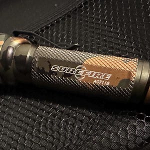 Puede incluir: Una linterna SureFire con un patr&oacute;n de camuflaje en tonos verdes, marrones y negros. La linterna tiene un agarre texturizado y un clip met&aacute;lico. El texto "SUREFIRE A07118" est&aacute; impreso en el lateral.