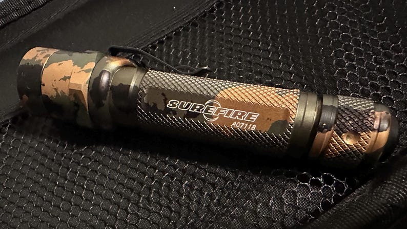 Puede incluir: Una linterna SureFire con un patr&oacute;n de camuflaje en tonos verdes, marrones y negros. La linterna tiene un agarre texturizado y un clip met&aacute;lico. El texto "SUREFIRE A07118" est&aacute; impreso en el lateral.