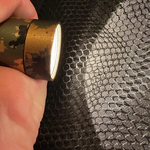 Puede incluir: Una linterna con estampado de camuflaje y bisel dorado emite un haz de luz brillante. La linterna se sostiene sobre un fondo oscuro y texturizado, creando un contraste entre la luz y la sombra.