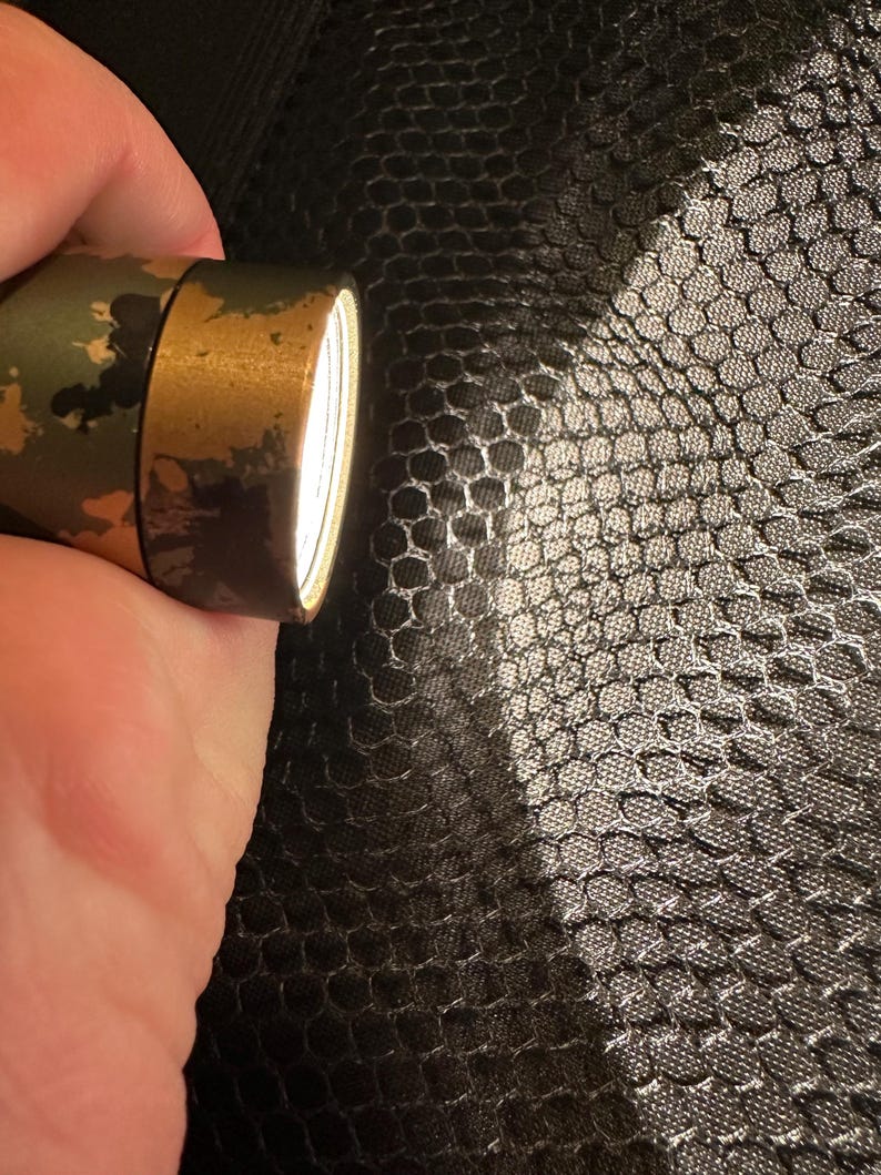 Puede incluir: Una linterna con estampado de camuflaje y bisel dorado emite un haz de luz brillante. La linterna se sostiene sobre un fondo oscuro y texturizado, creando un contraste entre la luz y la sombra.