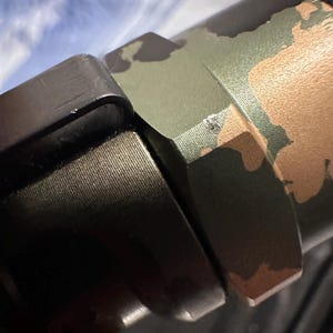 Puede incluir: Primer plano de una secci&oacute;n de un arma de fuego con un patr&oacute;n de camuflaje en tonos verdes, marrones y beige. La imagen muestra los componentes met&aacute;licos del arma de fuego, incluido un agarre negro texturizado y una pieza de conexi&oacute;n.