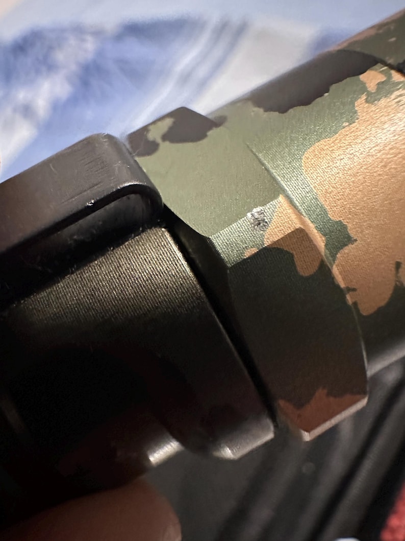 Puede incluir: Primer plano de una secci&oacute;n de un arma de fuego con un patr&oacute;n de camuflaje en tonos verdes, marrones y beige. La imagen muestra los componentes met&aacute;licos del arma de fuego, incluido un agarre negro texturizado y una pieza de conexi&oacute;n.