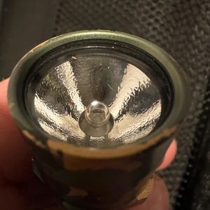 Puede incluir: Primer plano de una linterna con un patr&oacute;n de camuflaje en el exterior. La linterna tiene una lente transparente y una peque&ntilde;a bombilla en el centro. El borde met&aacute;lico alrededor de la lente es verde.