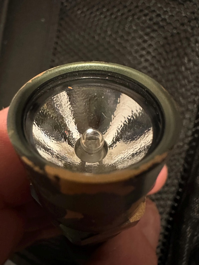 Puede incluir: Primer plano de una linterna con un patr&oacute;n de camuflaje en el exterior. La linterna tiene una lente transparente y una peque&ntilde;a bombilla en el centro. El borde met&aacute;lico alrededor de la lente es verde.