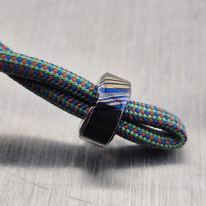 May include: A black ring with blue and yellow stripes is shown on a colorful climbing rope. The ring has a geometric design and is made of a dark material. The rope is green, red, and blue.