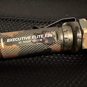 Puede incluir: Una linterna con estampado de camuflaje con el texto "EXECUTIVE ELITE E2e" y "US PATENT 6222138". La linterna tiene un agarre texturizado y un clip met&aacute;lico. La combinaci&oacute;n de colores incluye tonos de verde, marr&oacute;n y negro.