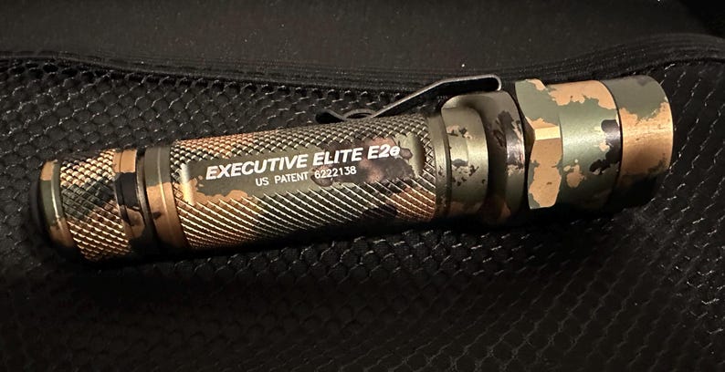 Puede incluir: Una linterna con estampado de camuflaje con el texto "EXECUTIVE ELITE E2e" y "US PATENT 6222138". La linterna tiene un agarre texturizado y un clip met&aacute;lico. La combinaci&oacute;n de colores incluye tonos de verde, marr&oacute;n y negro.