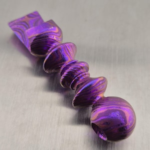 May include: A metallic object featuring a series of stacked, rounded, and spiraled segments with a vibrant purple and copper-toned swirling pattern. The piece has a polished, reflective finish and a distinct layered texture throughout its entire length.