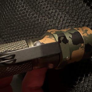Puede incluir: Primer plano de una linterna t&aacute;ctica con un patr&oacute;n de camuflaje en verde, marr&oacute;n y negro. La linterna tiene un agarre texturizado y un clip de bolsillo negro. El fondo es una superficie oscura y texturizada.