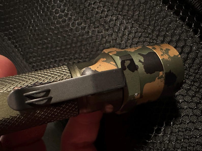 Puede incluir: Primer plano de una linterna t&aacute;ctica con un patr&oacute;n de camuflaje en verde, marr&oacute;n y negro. La linterna tiene un agarre texturizado y un clip de bolsillo negro. El fondo es una superficie oscura y texturizada.