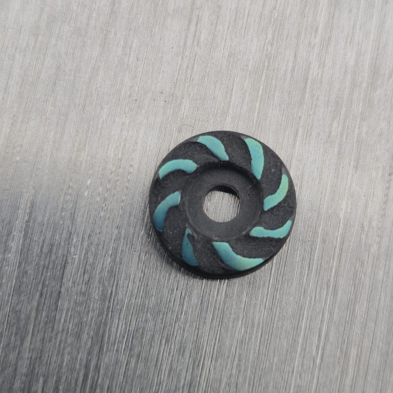 May include: A small, circular, black object with a central hole and teal-coloured curved accents. The teal accents are evenly spaced around the outer edge of the object, creating a spiral pattern. The object is set against a grey background.