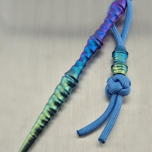 Op de afbeelding: Een veelkleurig metalen gereedschap met een getextureerde handgreep en een puntige punt. De tool gaat over van groen naar blauw naar paars. Een blauw koord met een decoratieve knoop is aan het gereedschap bevestigd.