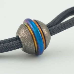 May include: A textured metal bead with a vibrant blue and gold center section, strung on a dark gray cord. The bead has a unique, iridescent finish, creating a striking visual contrast against the cord.