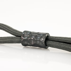 Peut inclure: Un cordon gris en paracorde avec une perle en métal noir texturée.