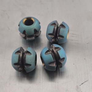 May include: Four small, metallic beads with a teal and gold finish. Each bead has a unique design with cut-out patterns and a central hole. The beads are arranged on a silver surface.