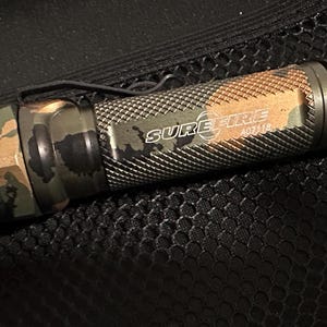 Puede incluir: Una linterna SureFire AD11B con un patr&oacute;n de camuflaje. La linterna tiene un clip de bolsillo negro y el logotipo de SureFire es visible. El cuerpo de la linterna es una mezcla de verde, marr&oacute;n y negro.