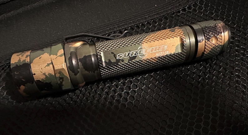Puede incluir: Una linterna SureFire AD11B con un patr&oacute;n de camuflaje. La linterna tiene un clip de bolsillo negro y el logotipo de SureFire es visible. El cuerpo de la linterna es una mezcla de verde, marr&oacute;n y negro.