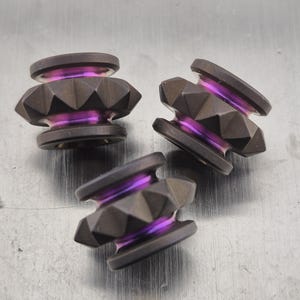 Puede incluir: Tres cuentas de metal gris oscuro con un diseño geométrico y detalles morados. Las cuentas tienen un patrón facetado en forma de diamante y son de forma cilíndrica. El color morado es visible en el centro de las cuentas.
