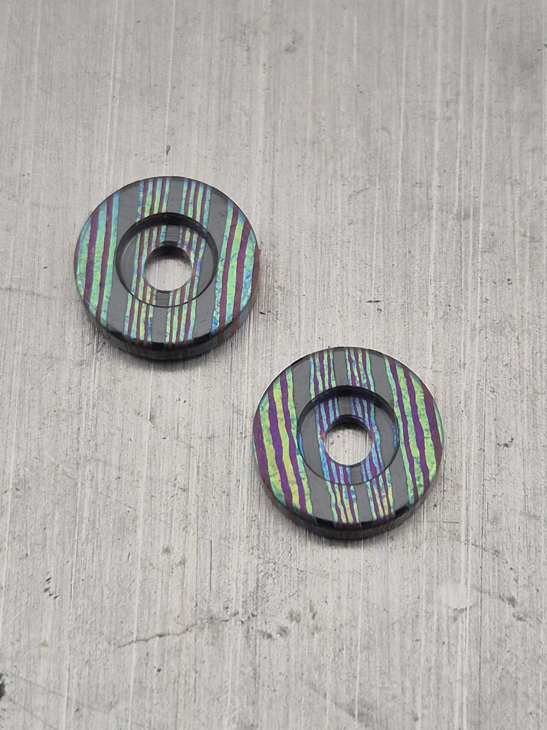 May include: Two circular objects with a central hole. The objects have a dark base color with vertical stripes in shades of purple, green, and yellow. The objects are on a gray surface.