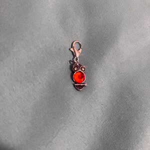 May include: A silver charm with a red jeweled owl design. The charm has a lobster clasp for attaching to a bracelet or necklace.