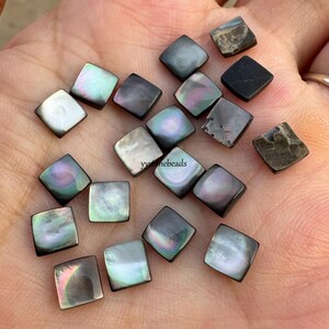 Puede incluir: Una colección de cuentas cuadradas de nácar iridiscente en varios tonos de gris, negro y blanco. Las cuentas tienen una superficie pulida y reflectante, con toques de rosa, verde y azul. Algunas cuentas tienen un aspecto rugoso y texturizado.