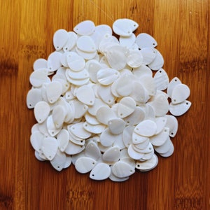 May include: A large pile of white pearlescent teardrop-shaped beads with a hole at the top. The beads are arranged in a random pattern and are all the same size.