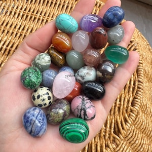 Op de afbeelding: Een verzameling van 20 getrommelde edelsteeneieren in verschillende kleuren en patronen, waaronder groen, roze, blauw, rood en zwart. De stenen zijn in een hand gerangschikt, met de tekst "yystonebeads" zichtbaar in het midden.
