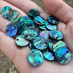 Könnte beinhalten: Eine Sammlung schillernder Abalone-Muschelkernperlen. Die runden Perlen haben ein Loch zum Auffädeln und zeigen eine Mischung aus blauen, grünen und violetten Farbtönen. Die Perlen werden in einer Hand gezeigt.