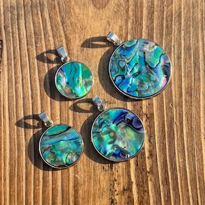 May include: Four round pendants with iridescent abalone shell in silver settings. The pendants vary in size, displaying blue, green, and purple colours. The pendants are presented on a wooden surface.