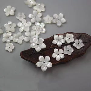 May include: White flower-shaped embellishments scattered on a gray surface, with some arranged on a dark brown, leaf-shaped wooden piece. The flowers have five petals and a small hole in the center, suggesting they are beads or decorative elements.