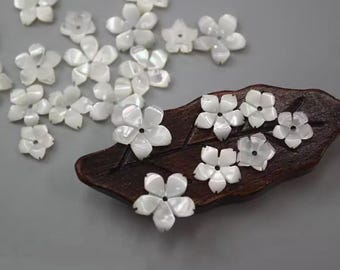 May include: White flower-shaped embellishments scattered on a gray surface, with some arranged on a dark brown, leaf-shaped wooden piece. The flowers have five petals and a small hole in the center, suggesting they are beads or decorative elements.