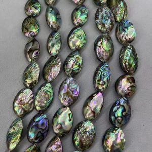 May include: Close-up of iridescent abalone shell beads. The oval-shaped beads display a range of colors, including green, purple, and blue, with a shimmering, textured surface. The beads are strung together, suggesting they are for jewelry making.