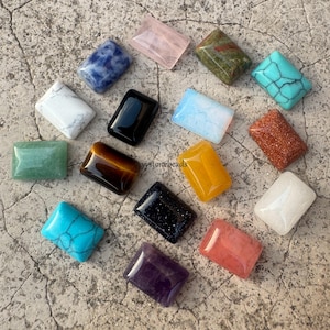 May include: A selection of rectangular gemstones in diverse colours, such as blue, pink, green, and gold. The gemstones are displayed on a textured, neutral-toned surface. The image presents a variety of natural stones.