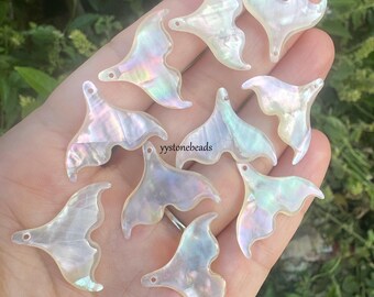 May include: A collection of iridescent white whale tail charms with a pearlescent finish, each featuring a small hole for attachment. The charms display a smooth, reflective surface with subtle rainbow hues. The text yystonebeads is visible.