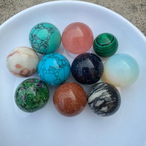 May include: Ten polished gemstone spheres in various colours and patterns, including turquoise, pink, green, white, brown, and black. The spheres are arranged in a circular pattern on a white plate.