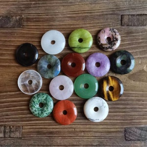 May include: Assortment of 15 donut-shaped gemstone pendants in various colors, including green, white, black, red, purple, and brown. The pendants are arranged in a circle on a wooden surface.