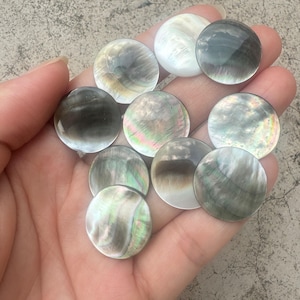 Puede incluir: Surtido de cabujones redondos de nácar iridiscente en tonos de negro, gris y blanco. Los cabujones son lisos y pulidos, con un brillo sutil.