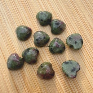 May include: Ten green and pink heart-shaped stones with black flecks. The stones are arranged on a wooden surface.