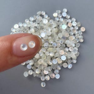 May include: A collection of small, round, iridescent moonstone beads. Two beads are held between a finger and thumb, showcasing their milky white and subtle rainbow hues. The beads are scattered on a gray surface, with the text "yystonebeads".