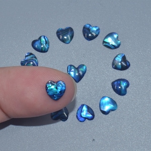 May include: Assortment of blue iridescent heart-shaped shell cabochons. The cabochons are arranged in a circle with one heart being held by a finger.