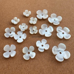 May include: A collection of white, iridescent mother-of-pearl beads shaped like four-petaled flowers. Each bead features a central hole for threading. The beads vary in size and display a smooth, glossy finish with natural pearlescent luster.