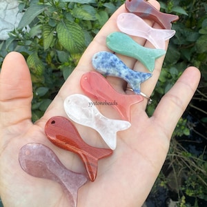 Könnte beinhalten: Eine Sammlung von acht polierten Edelsteinanhängern in Fischform in verschiedenen Farben wie Rot, Weiß, Hellrosa, Grün und Blau, jeweils mit einem kleinen Bohrloch am Kopf und dem sichtbaren Text yystonebeads versehen.