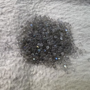 Puede incluir: Un montón grande de cuentas redondas de piedras preciosas transparentes e iridiscentes. Las cuentas están esparcidas sobre una superficie blanca y peluda.